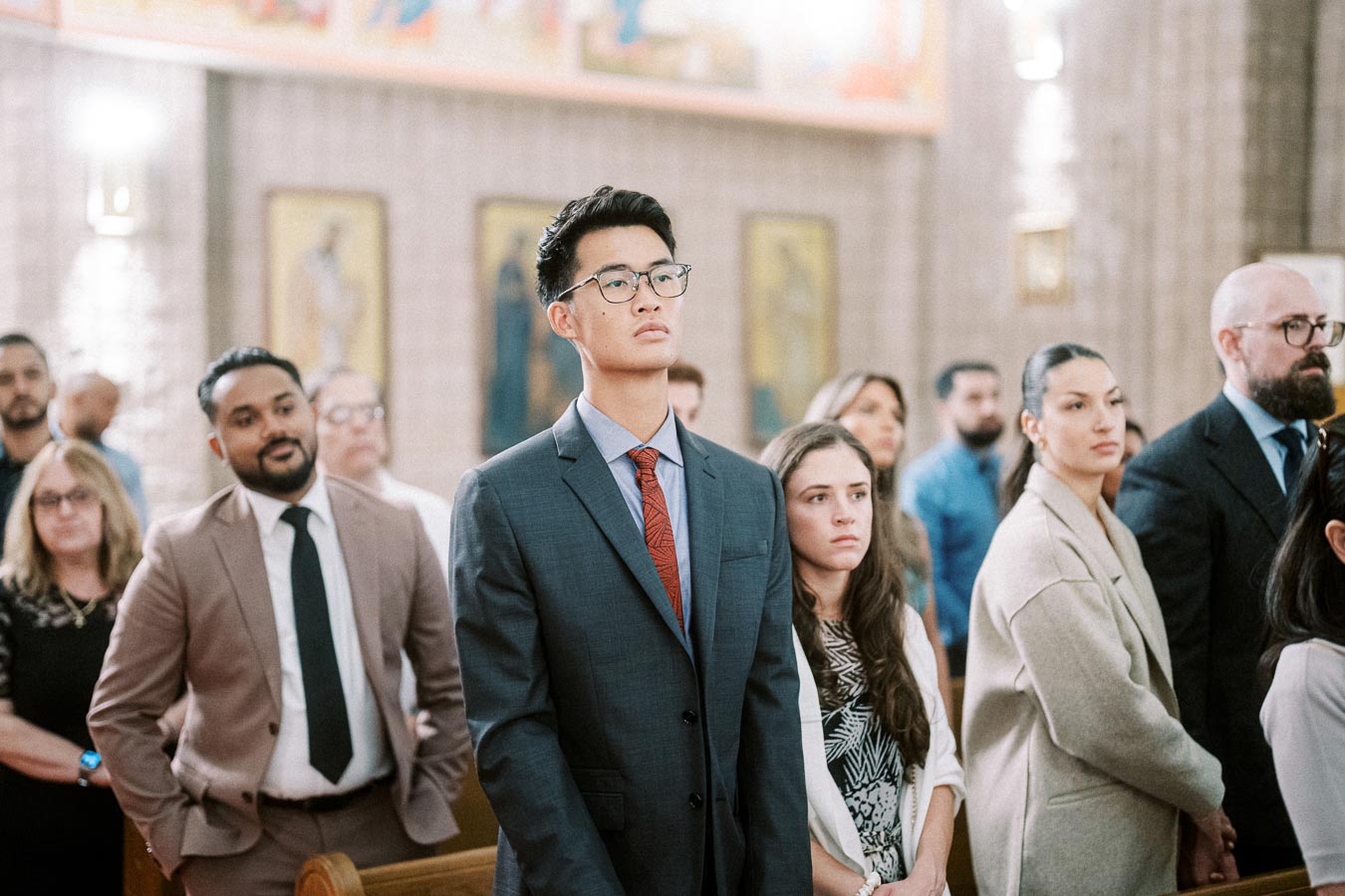 A diverse group of people standing attentively in a church setting, wearing formal attire, with artwork and religious icons