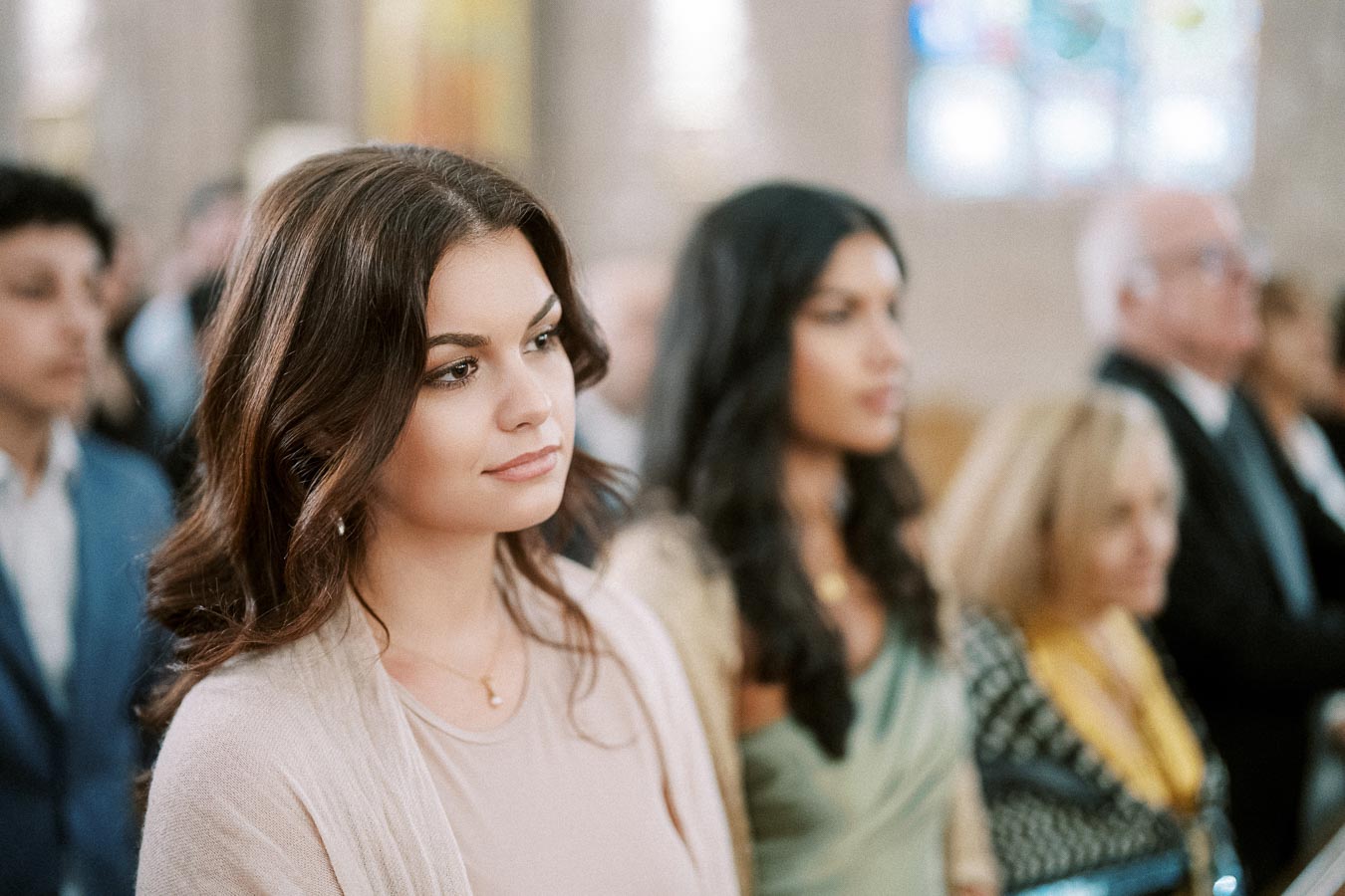 Young woman with long brown hair attending an event in a softly lit venue, surrounded by a diverse group of people in formal