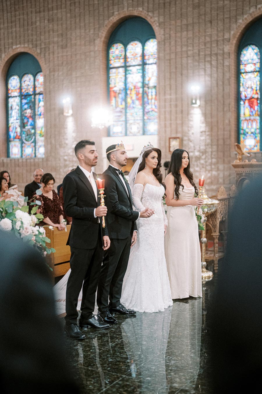 A bride and groom wearing crowns stand together with their wedding party holding candles in a church adorned with stained