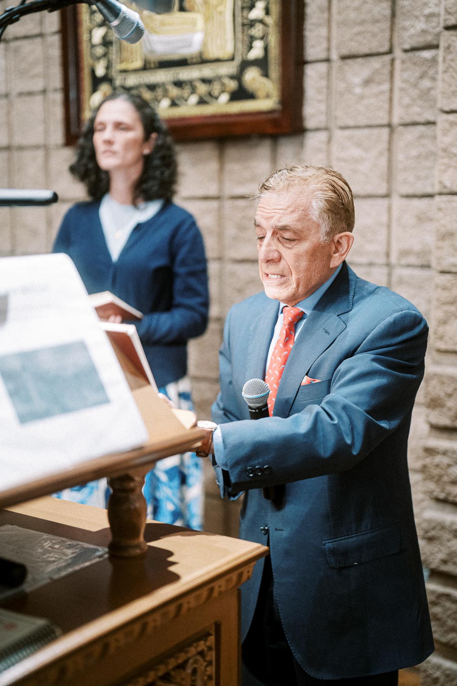 Man in a blue suit speaking into a microphone at a podium with a woman in the background, both in a formal setting.