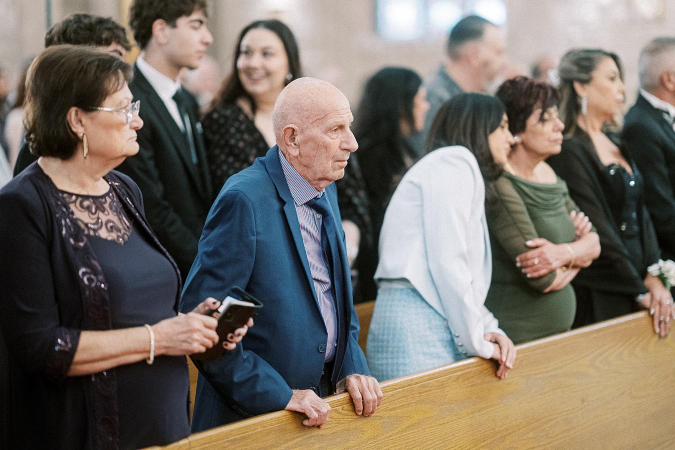 A group of people, including an elderly man in a blue suit, stand attentively in a church pew during a ceremony.