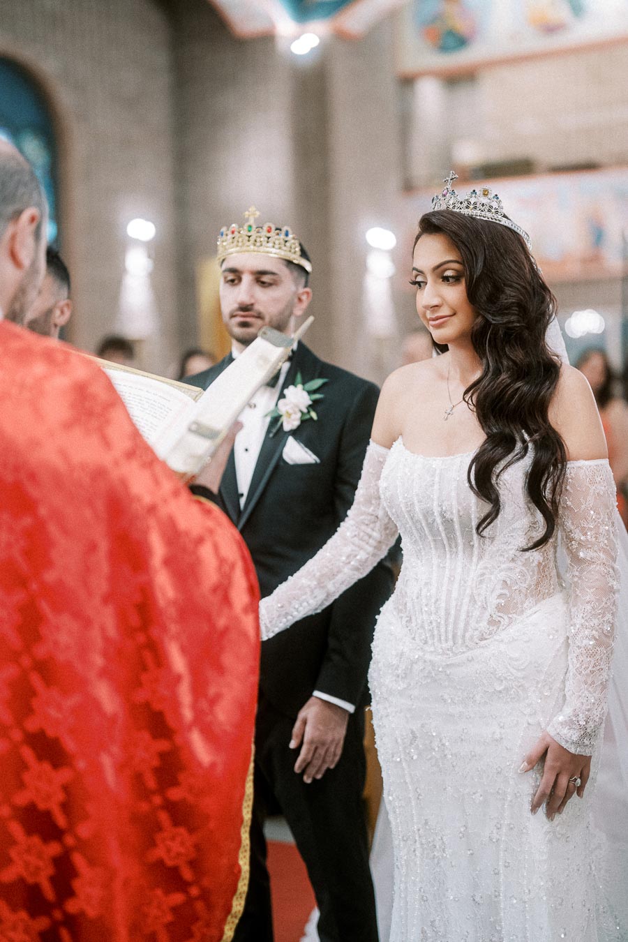 A bride and groom wearing crowns during a traditional wedding ceremony, facing a priest in red robes who is holding a book