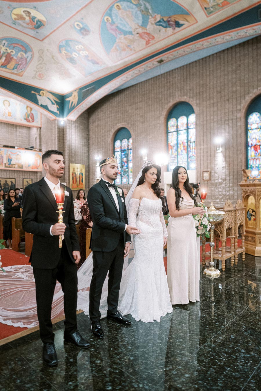 A bride and groom wearing crowns stand during a traditional wedding ceremony inside an ornately decorated church with