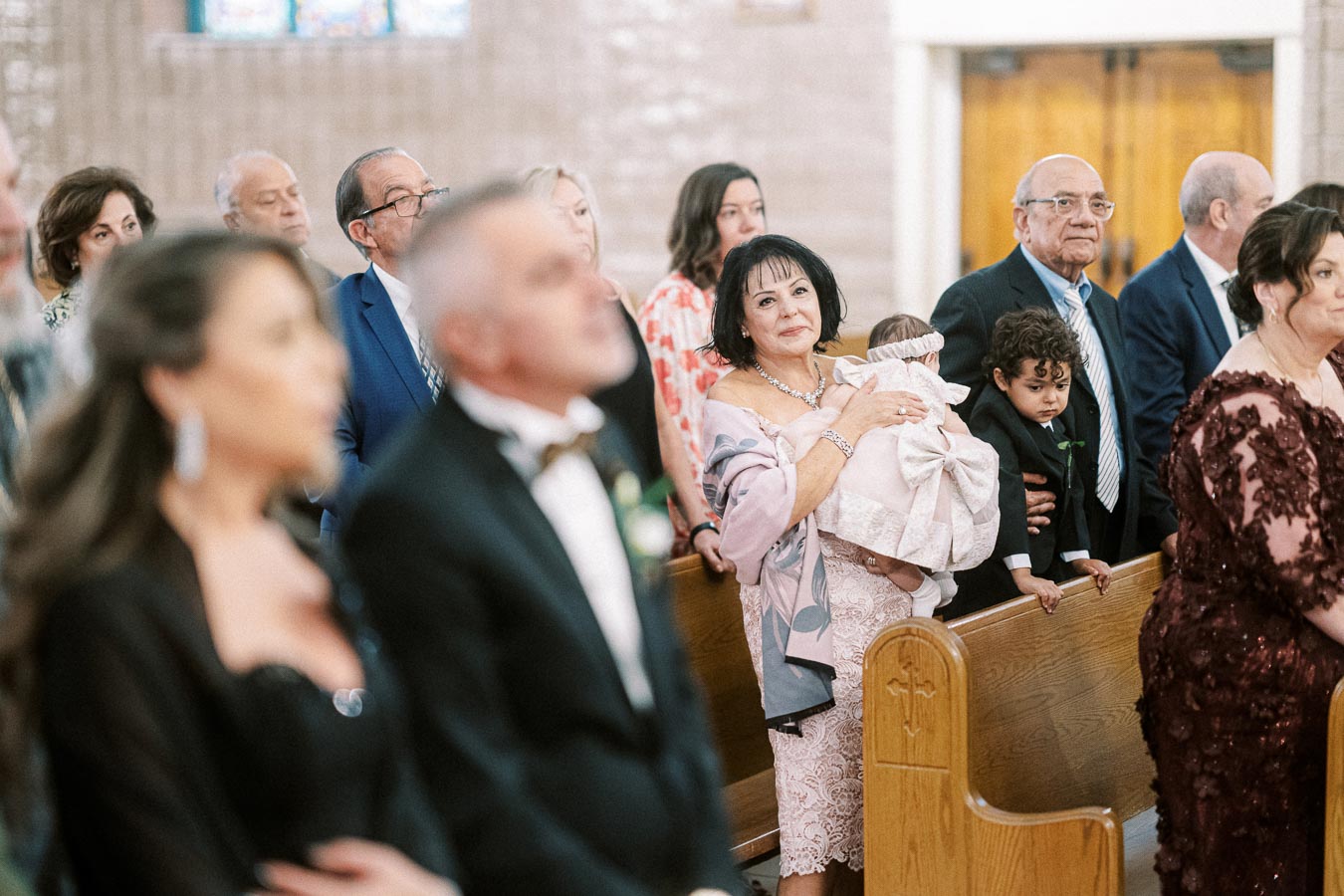 A group of well-dressed individuals attending a formal event in a church setting, with a woman holding a baby wrapped in a