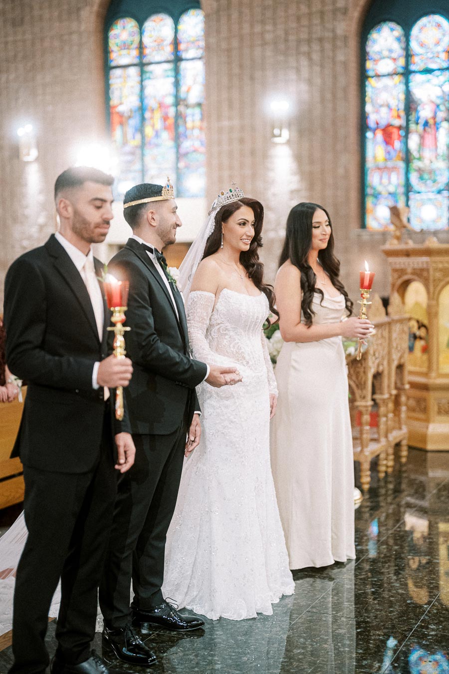 Ceremonial wedding scene inside a church with a bride and groom wearing crowns, standing together with attendants holding
