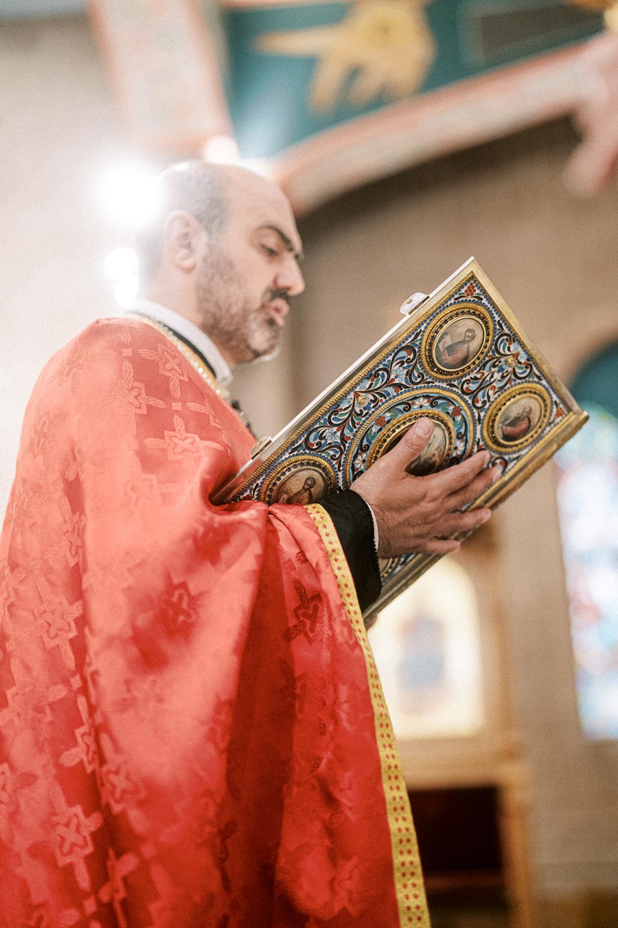 Priest in ornate red robe reading a decorative religious book inside a church.