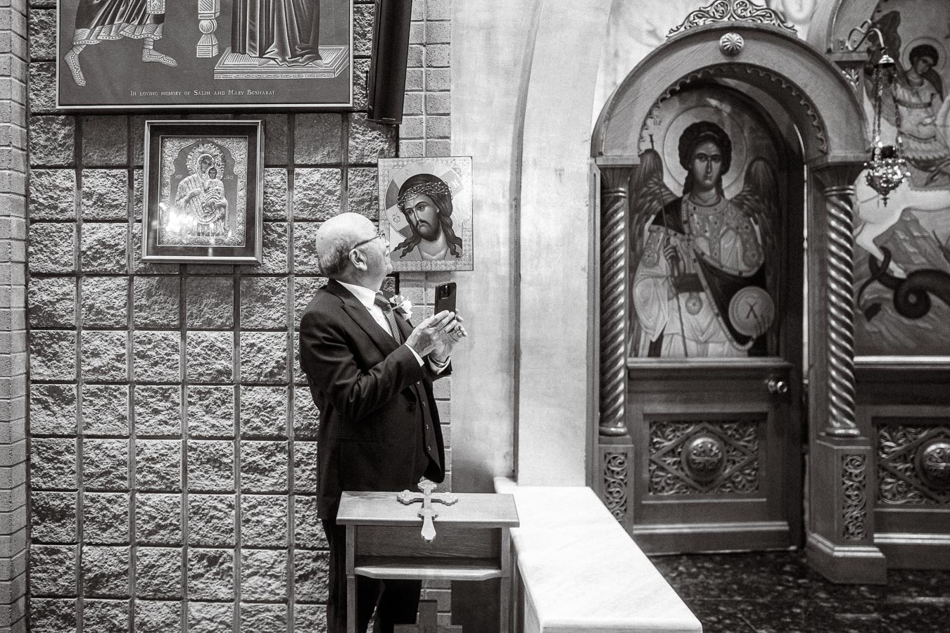 Elderly man in a suit holding a smartphone, standing in a church beside religious iconography, featuring framed images of