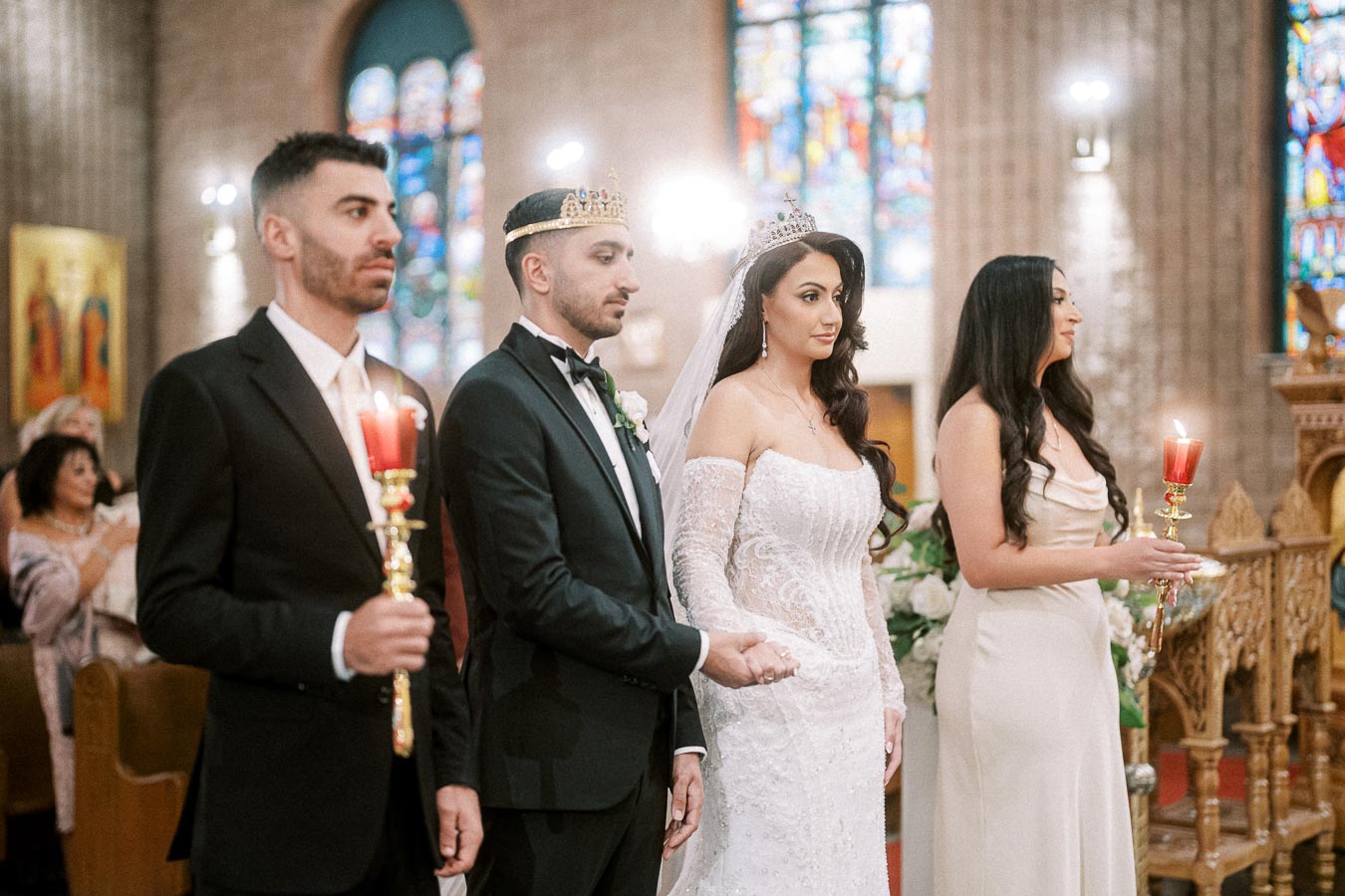 A bridal couple and their attendants stand in a beautifully decorated church, wearing formal attire and holding candles,