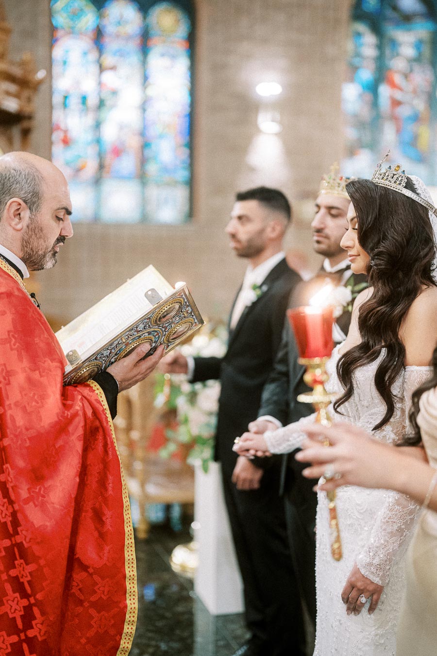 A priest in a red robe reads from a ceremonial book during a traditional wedding ceremony inside a church with stained glass