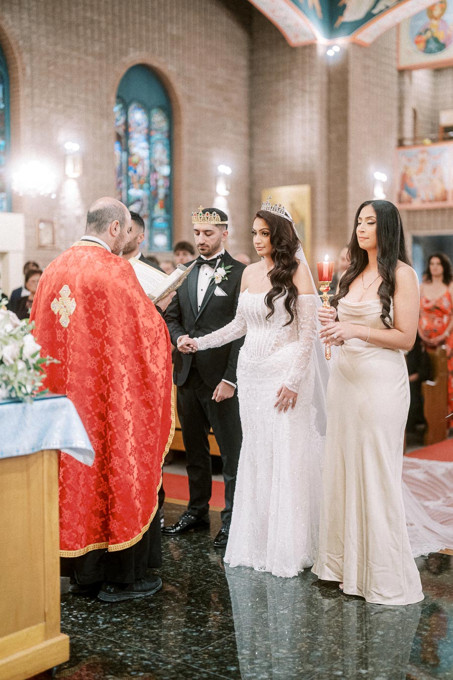 A wedding ceremony in a church with a priest conducting the service. The bride, in a white gown and crown, holds hands with