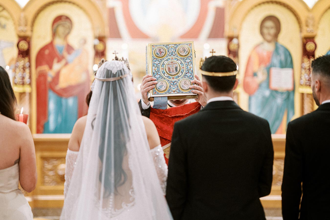 Orthodox wedding ceremony with bride and groom in front of a priest holding a decorated religious book, surrounded by