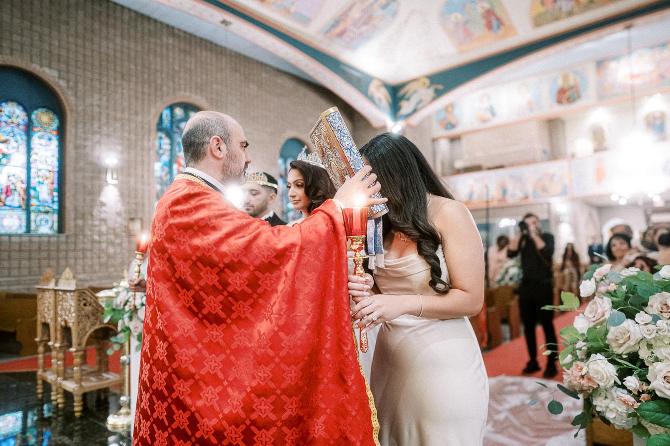 Ceremony in a decorated church with a priest in red robes, blessing a couple during a traditional wedding ritual, with