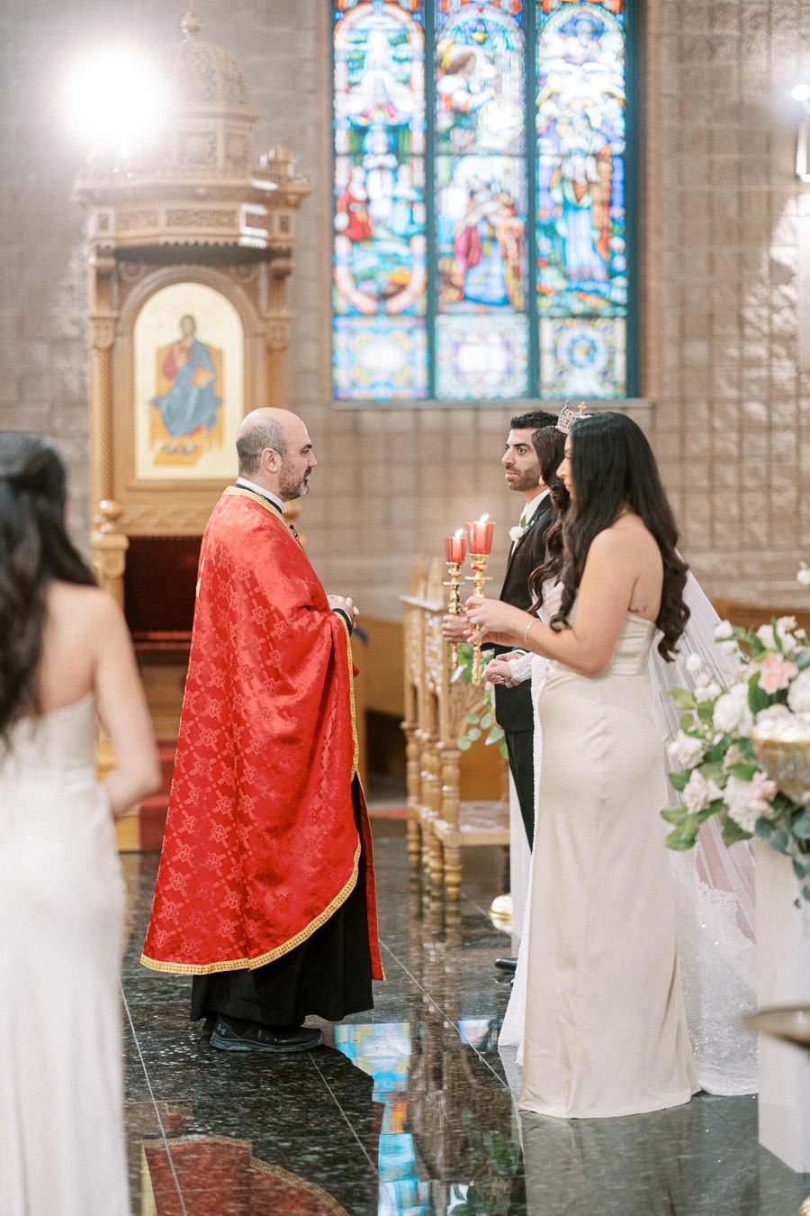 Couple during a traditional church wedding ceremony, with a priest in red robes, stained glass windows in the background,