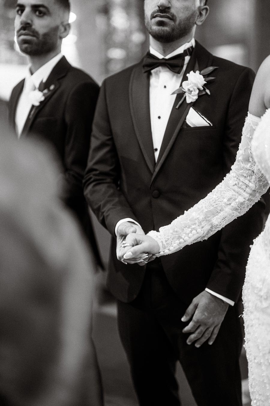 Black and white image of a wedding couple holding hands, the groom is in a tuxedo with a boutonnière, while the bride wears