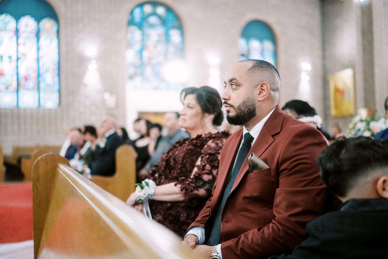 A man in a red suit attentively sits in a church pew, surrounded by other well-dressed attendees. Stained glass windows