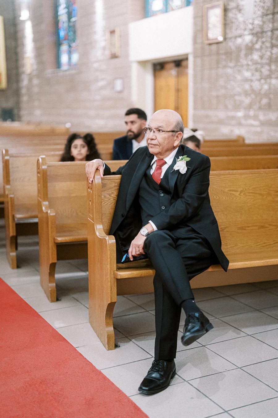 Elderly man wearing a suit with a flower boutonniere, seated in a church pew, attending a ceremony with others in the