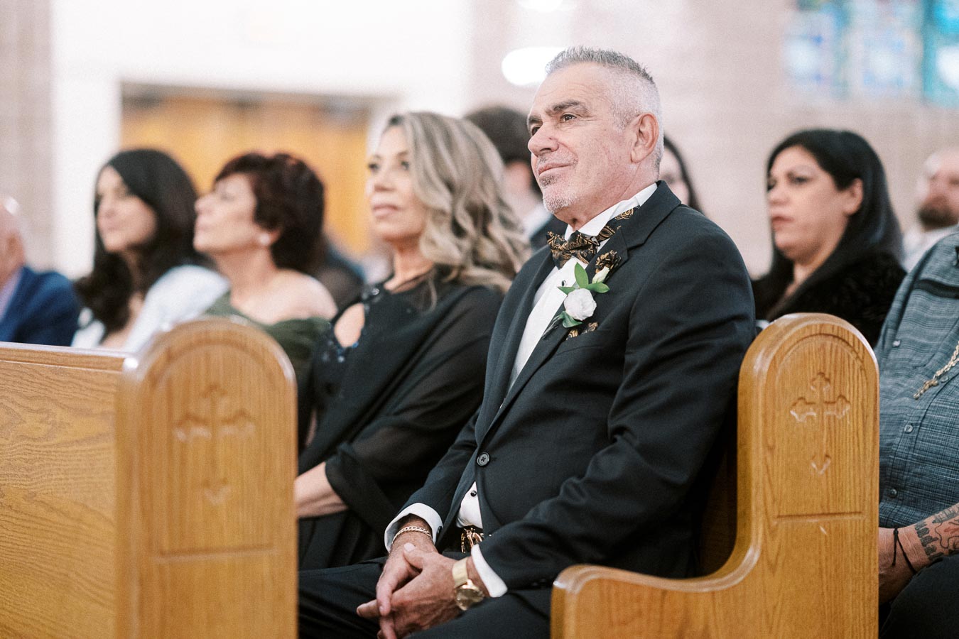 Elegant wedding guests seated in church pews, featuring a distinguished man in a suit with a boutonniere, attentively