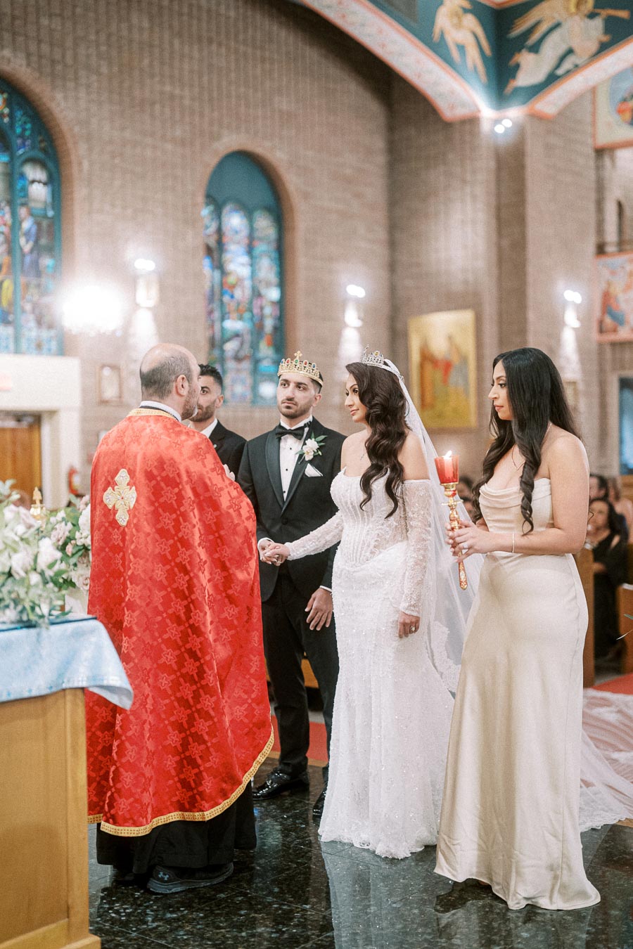 Elegant church wedding ceremony with bride and groom wearing crowns, holding hands in front of a priest in a red robe,