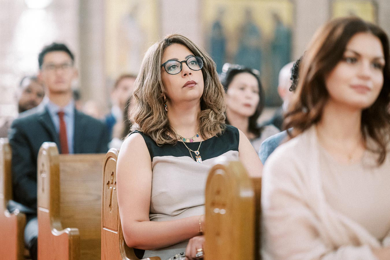A thoughtful woman with glasses sitting in a church pew, surrounded by other attendees, in a solemn and attentive atmosphere.