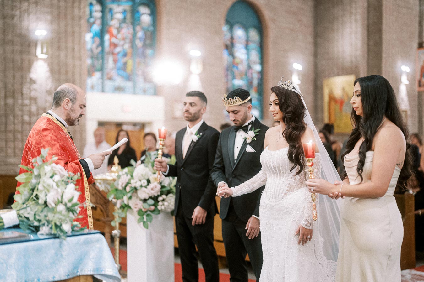 A priest officiates a traditional wedding ceremony inside a decorated church, with the bride and groom wearing crowns and