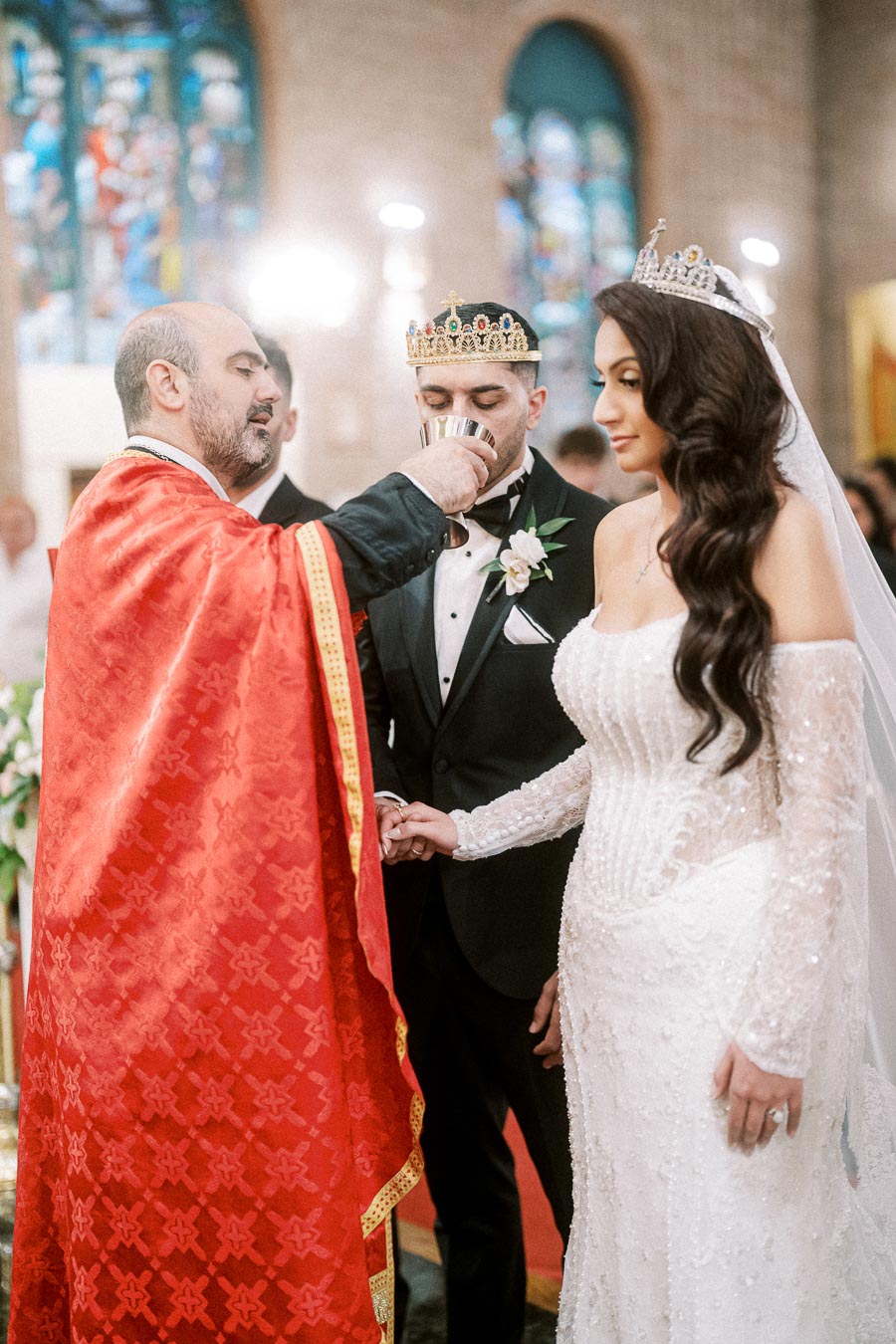 A couple wearing crowns is solemnly holding hands during a traditional wedding ceremony in a church, with a priest in a red