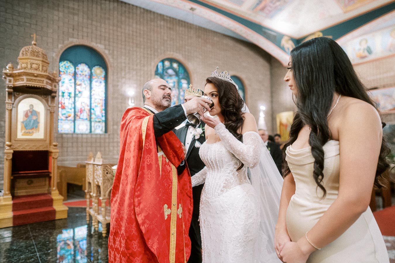 Bride and groom participate in a traditional wedding ceremony inside an ornate church, with a priest in red vestments