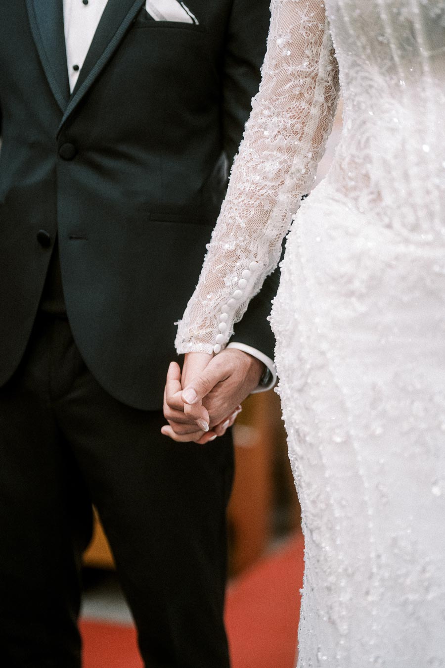 Bride and groom holding hands during their wedding ceremony, wearing an elegant white lace gown and a classic black tuxedo.