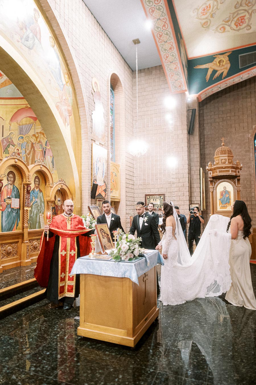 Orthodox wedding ceremony in a richly decorated church interior, featuring a priest in red robes, a couple in wedding