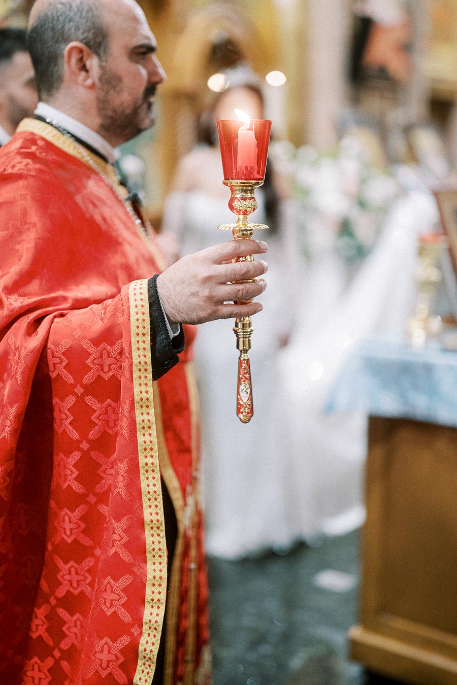 Clergyman in ornate red robe holding a lit candle during a religious ceremony in a church setting.