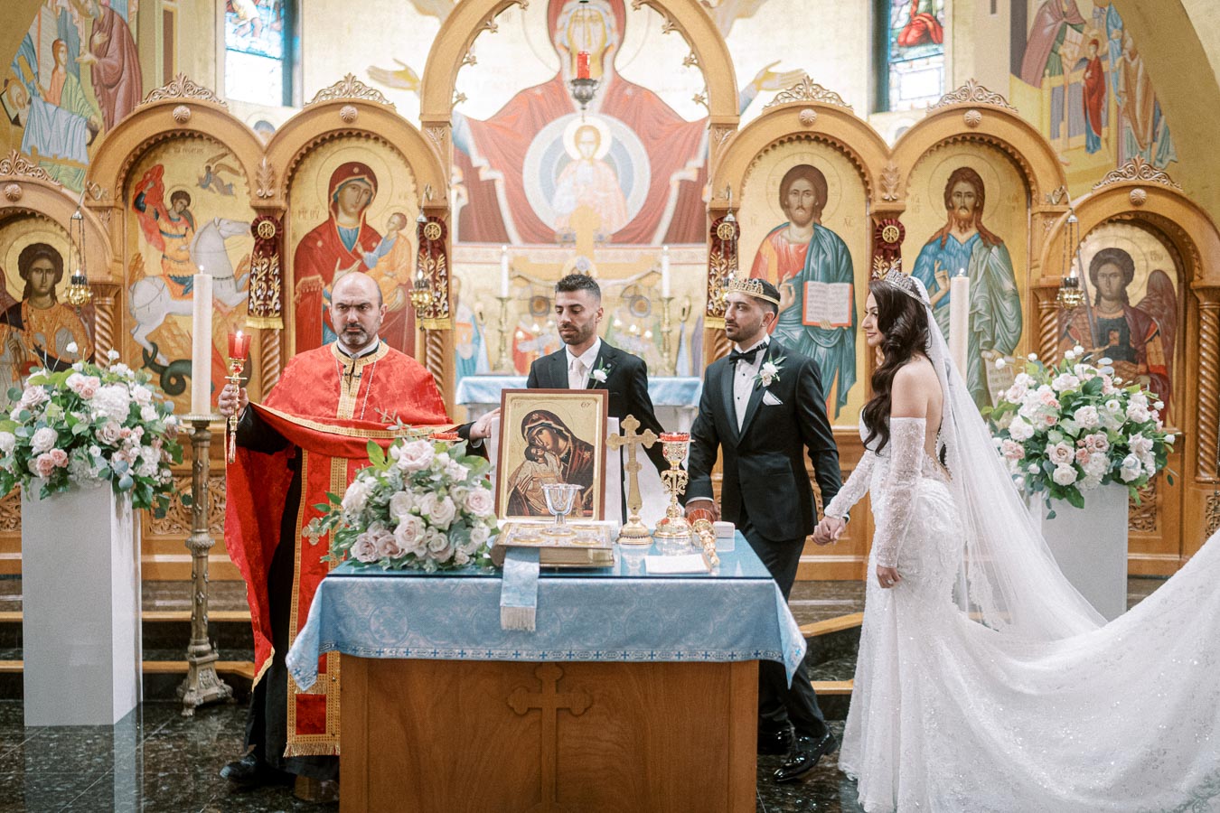 Orthodox wedding ceremony in a richly decorated church with icons in the background. A priest in red robes leads the