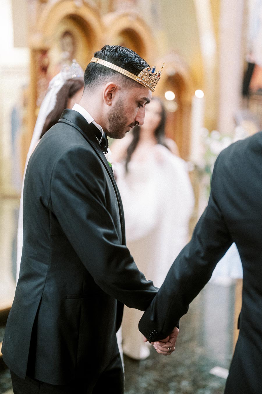 Man in a suit wearing a decorative crown, holding hands during a traditional ceremony.