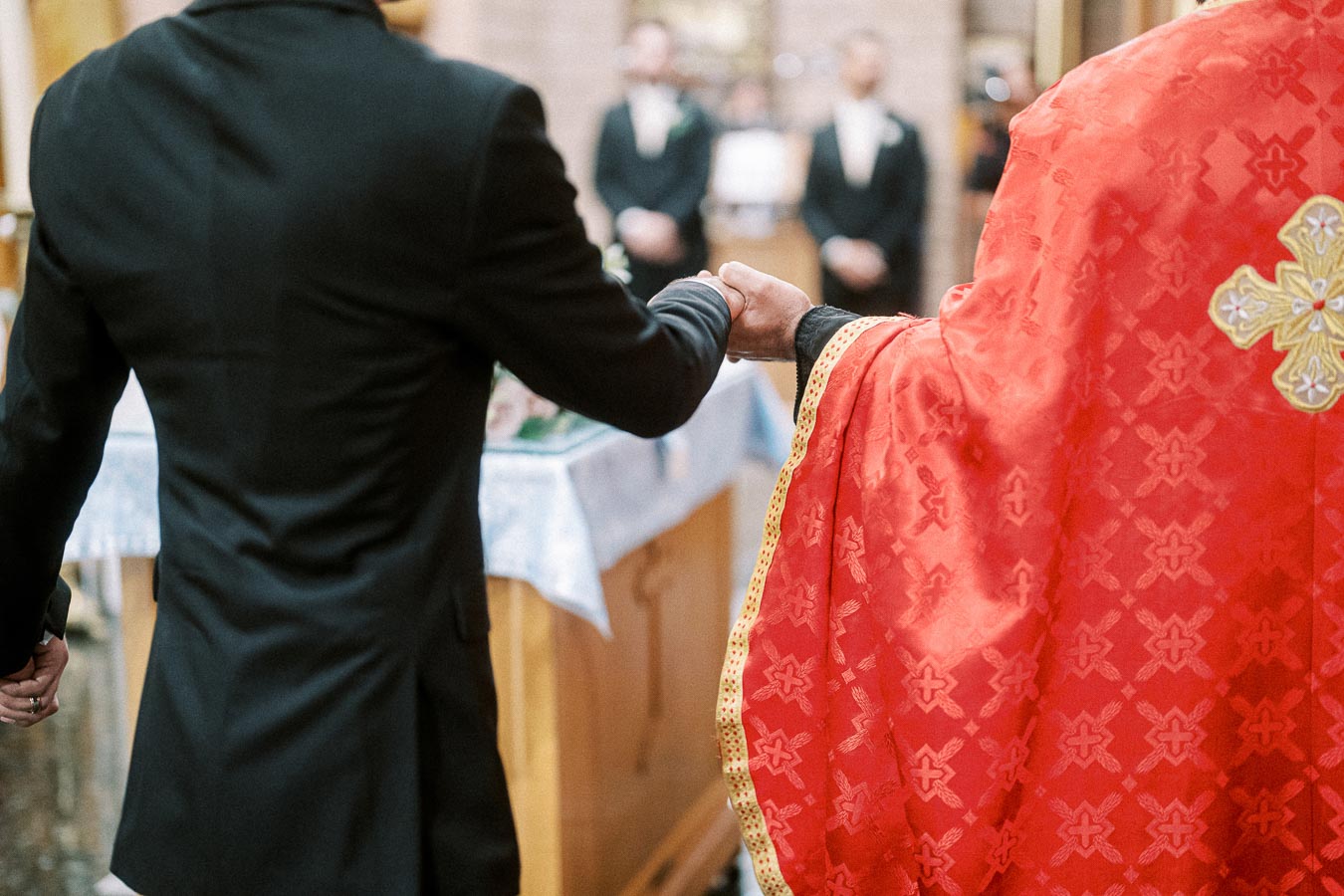 A priest in a red robe holds hands with a man in a black suit during a wedding ceremony inside a church, with blurred guests