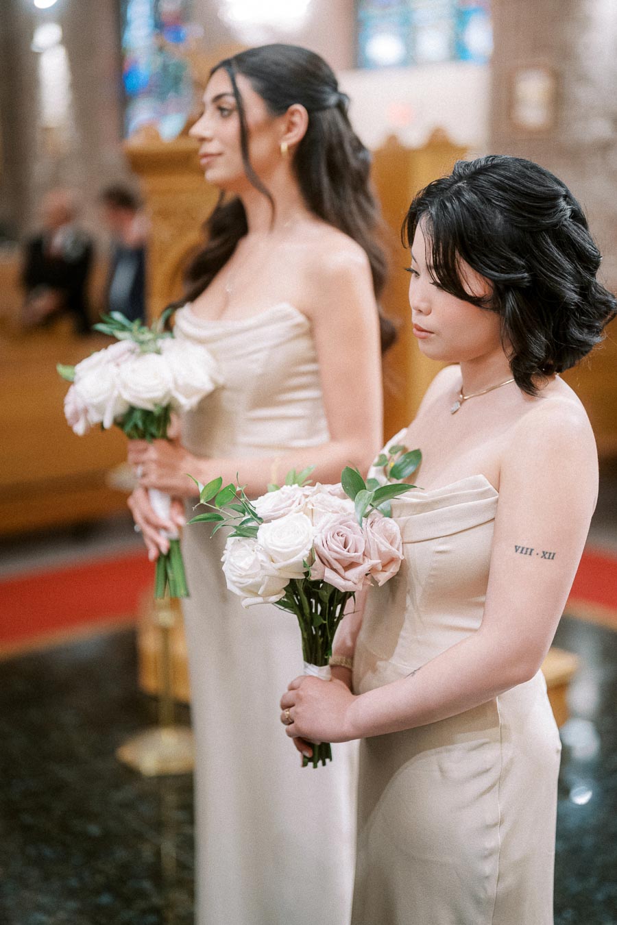 Two bridesmaids in elegant beige dresses holding bouquets of pale pink roses during a wedding ceremony in a church setting.