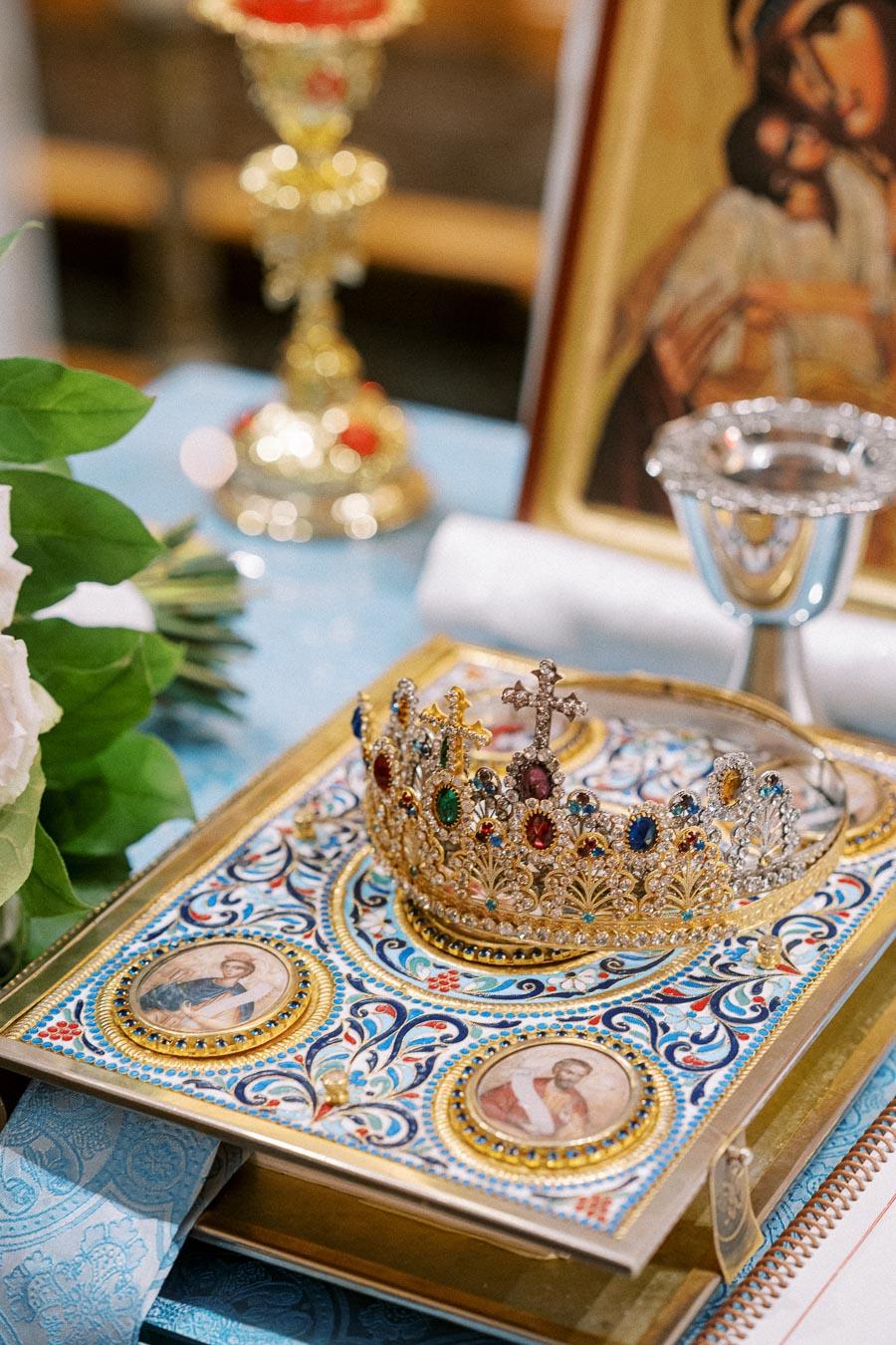Ornate crown adorned with colorful jewels resting on a richly decorated religious book, surrounded by a chalice