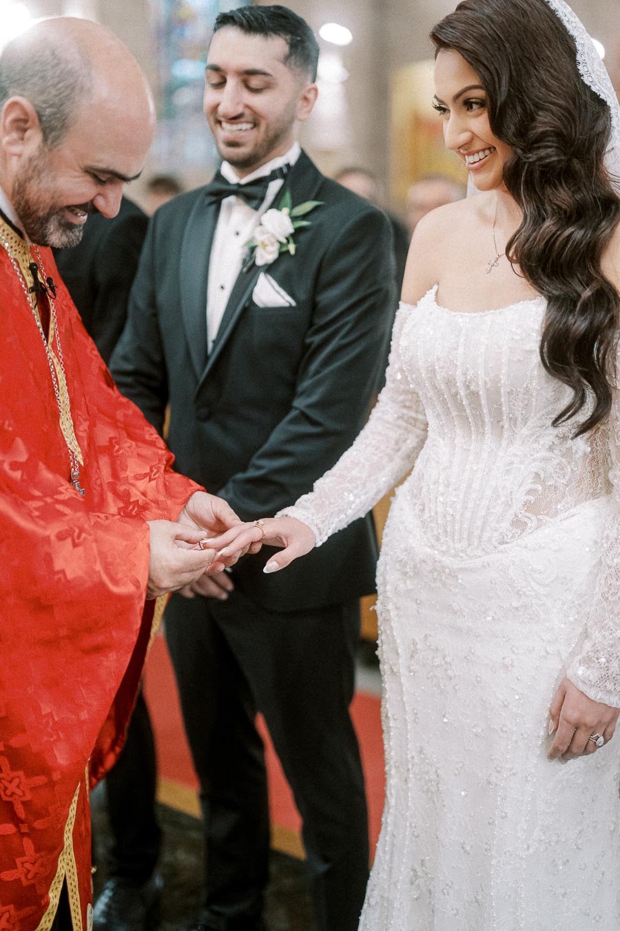 Wedding ceremony scene with a smiling bride wearing a white lace gown and a groom in a black tuxedo. An officiant in a red