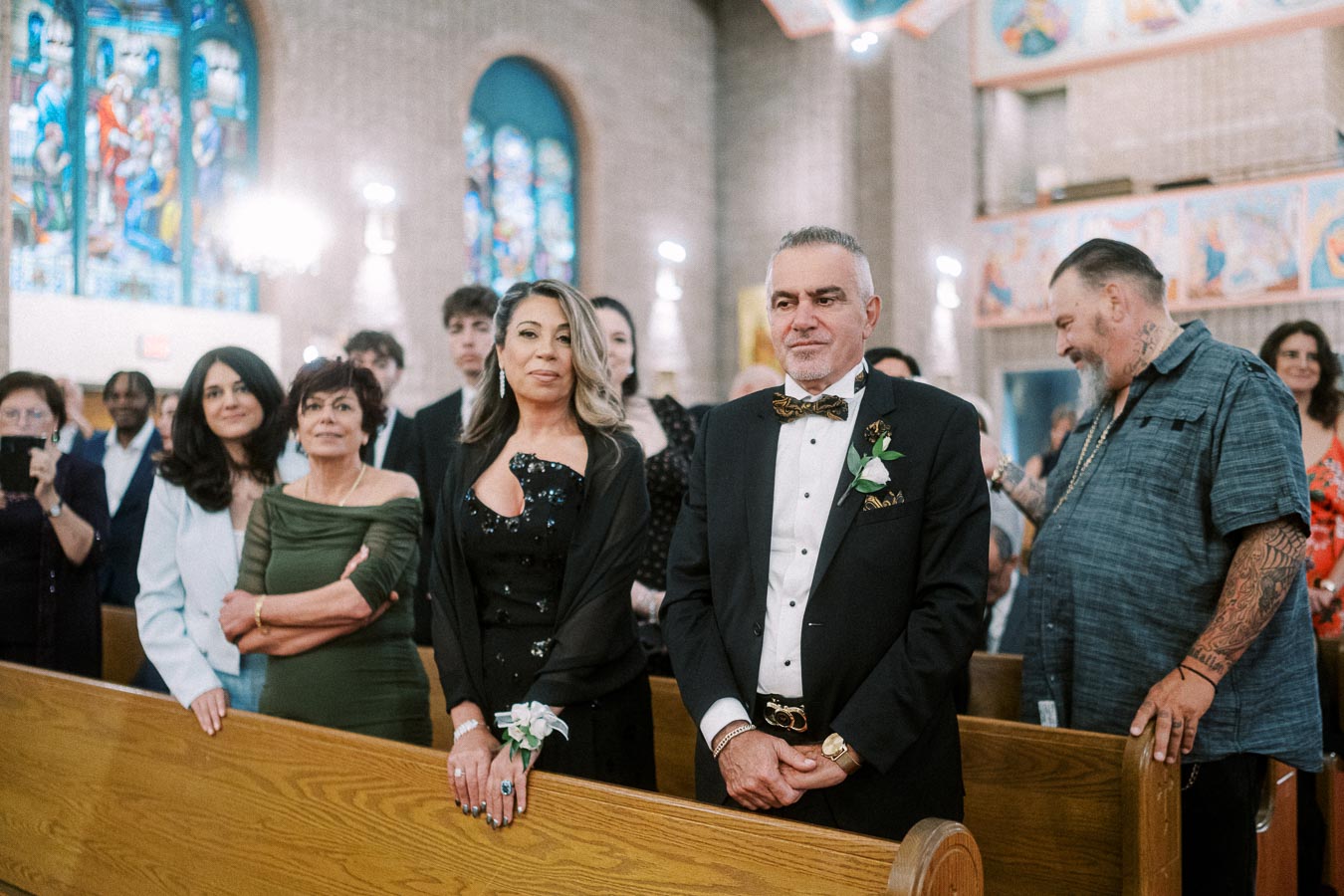 A group of people at a formal event in a church, including a well-dressed couple in the foreground with stained glass