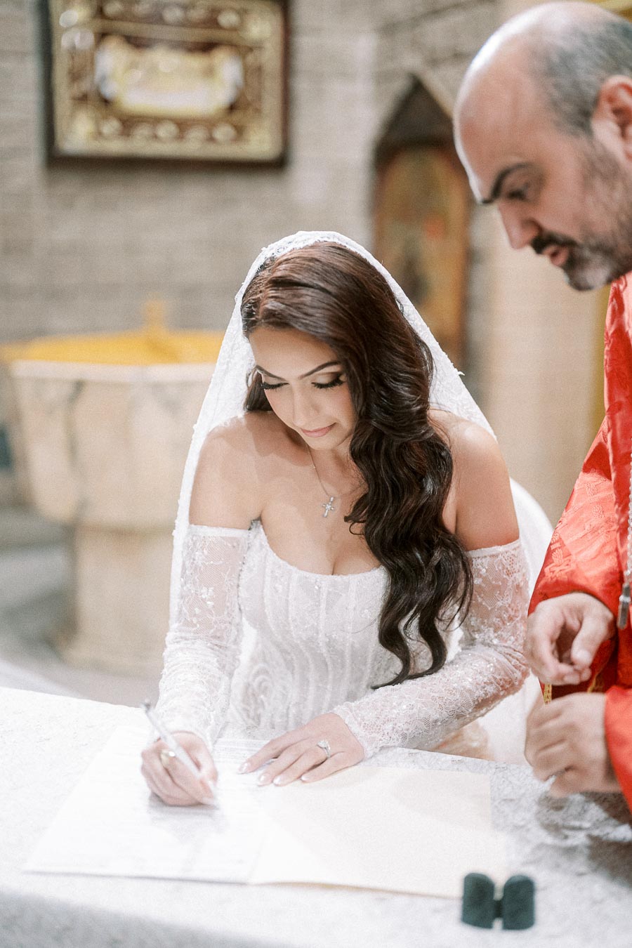 A bride in a lace wedding dress signs a document while a man in red looks on in a church setting, emphasizing the elegance