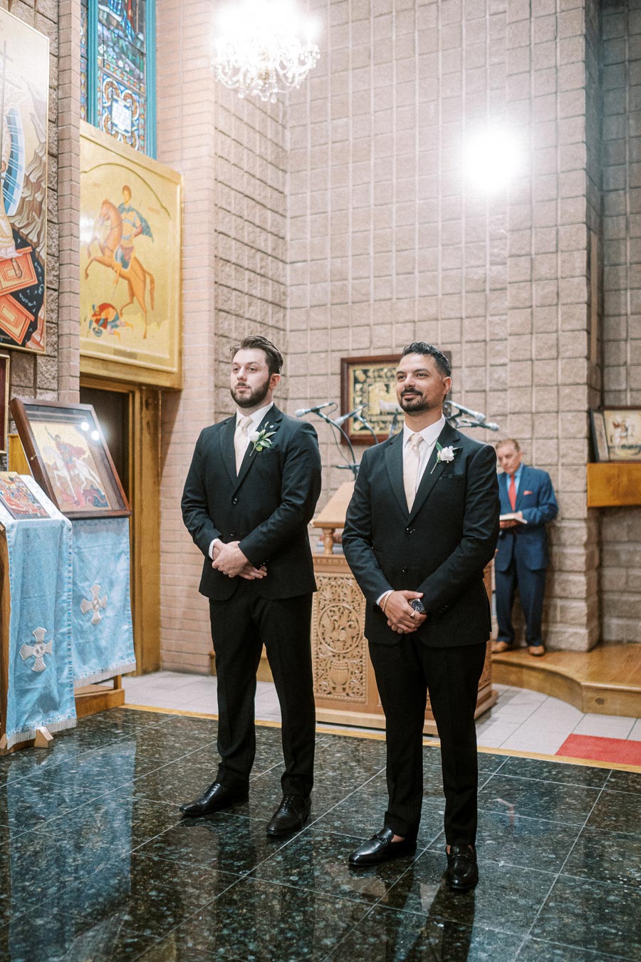Two groomsmen in black suits and white ties stand confidently in an elegantly decorated church, with vibrant religious