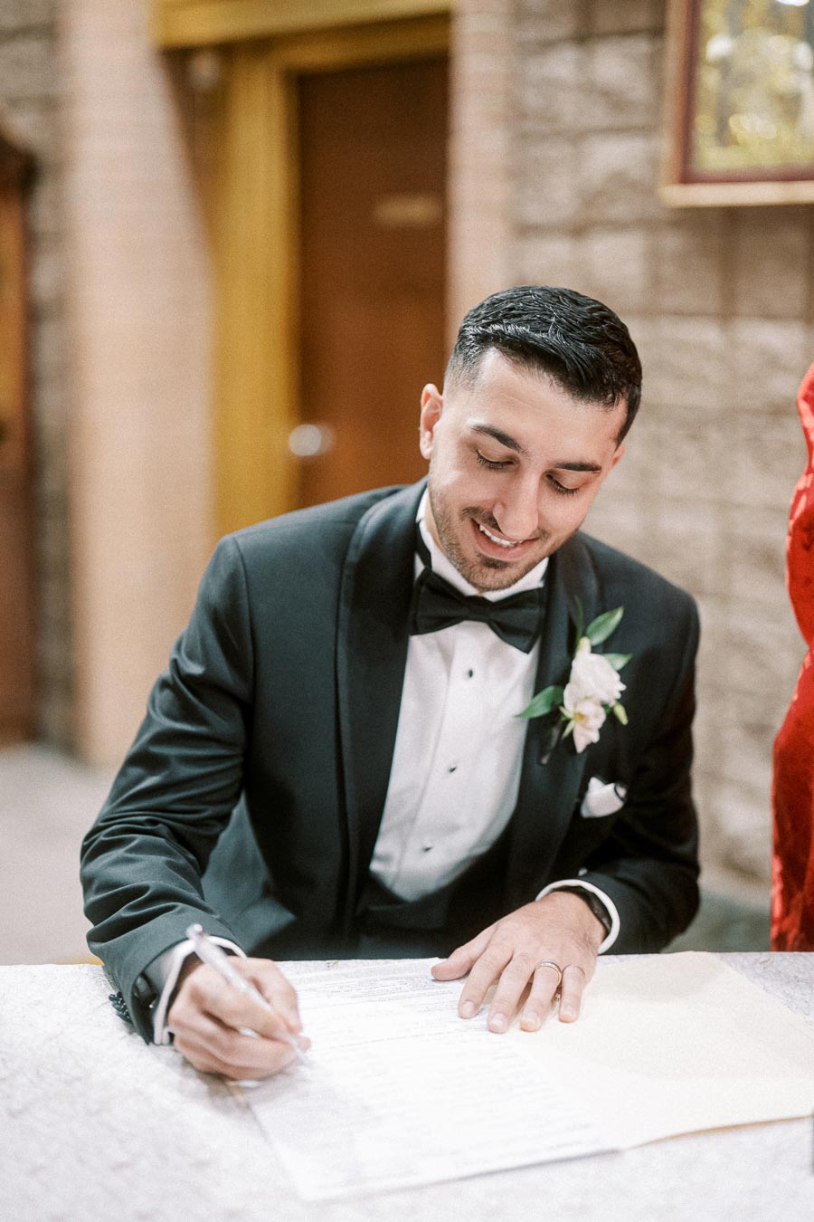 Man in black tuxedo with floral boutonniere signing wedding document at ceremony.