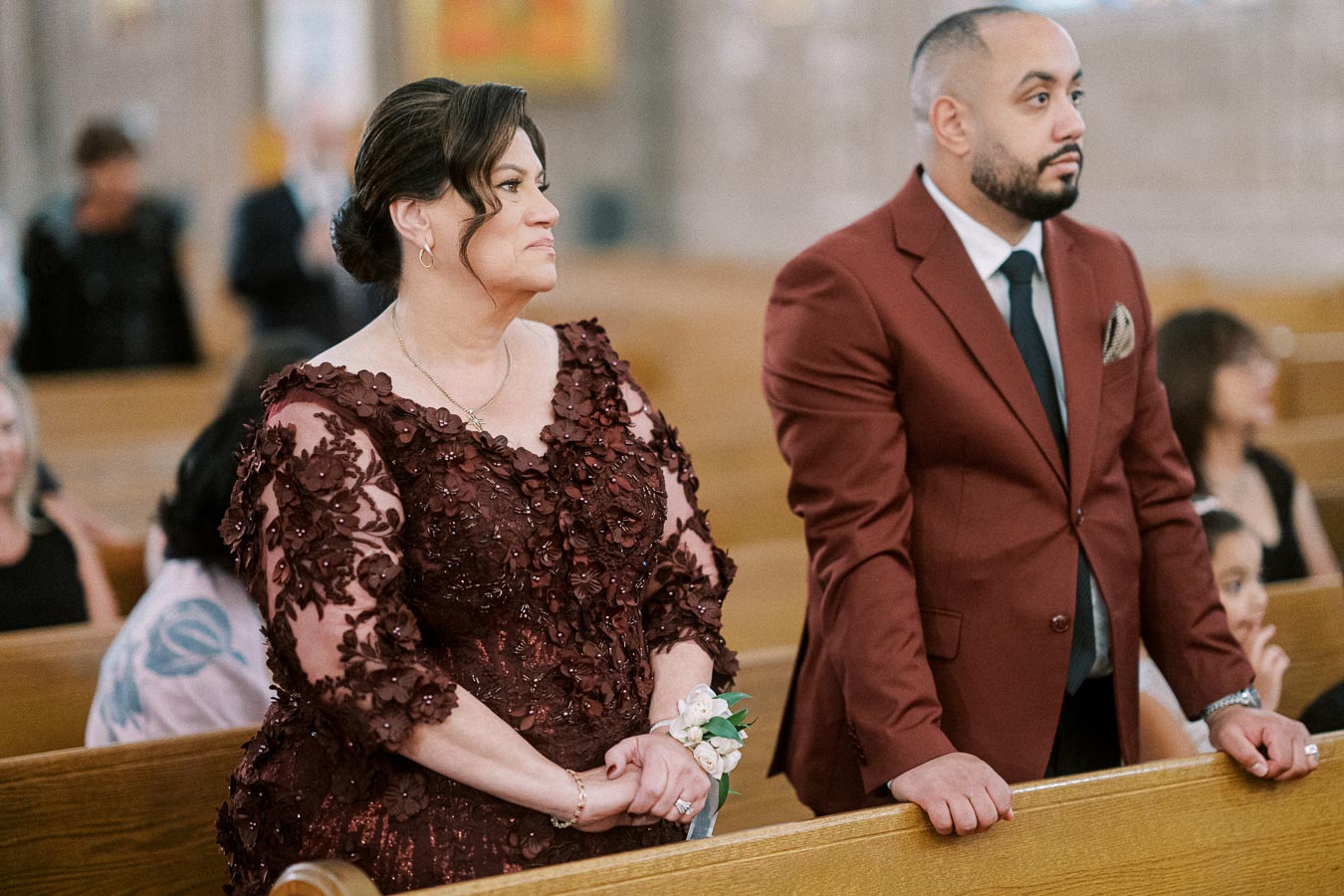 A man and woman dressed formally, standing in a church pew during a ceremony. The woman wears a floral burgundy dress with a