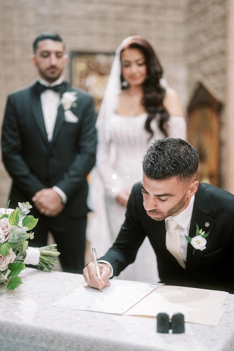 Groom signing a marriage license at a wedding ceremony with the bride and best man standing in the background, elegantly