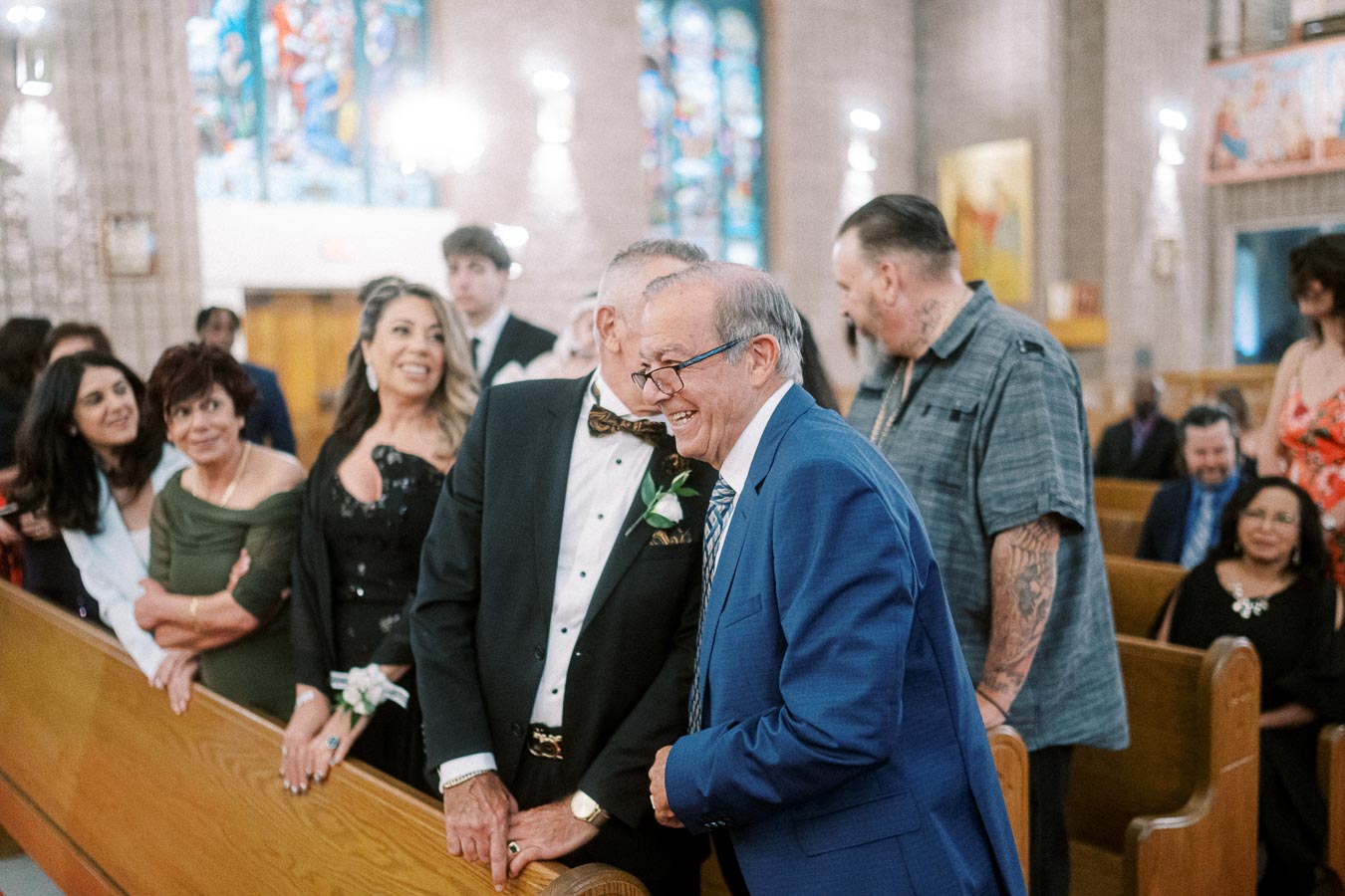 A joyful gathering of people dressed in formal attire, conversing and smiling inside a church with stained glass windows.