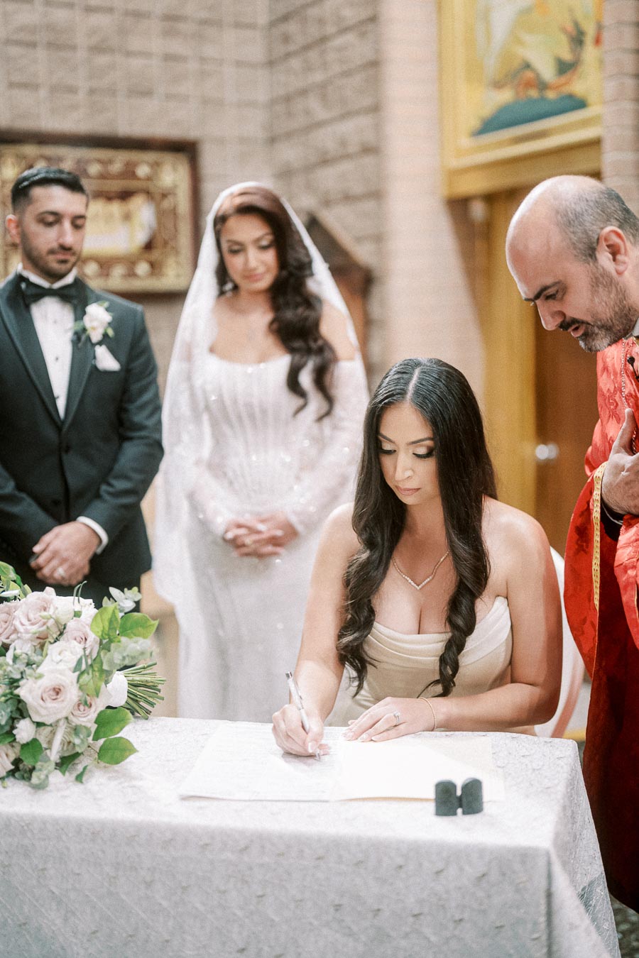 A bride and groom stand together as a woman signs a document during a wedding ceremony, with a priest in red overseeing the