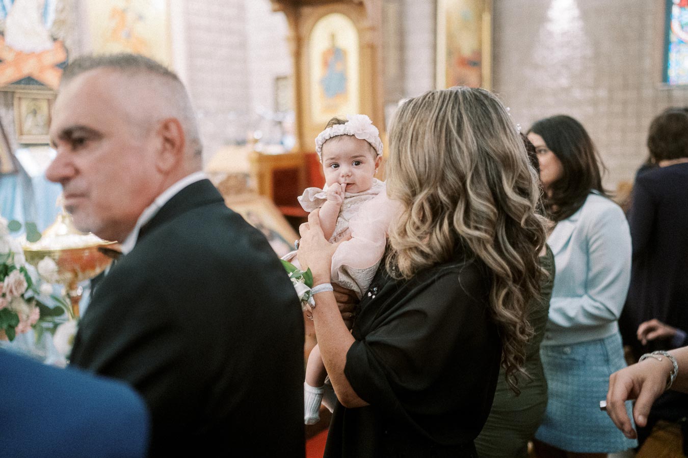 A baby wearing a white headband is held by a woman during a church event, surrounded by people and soft lighting.