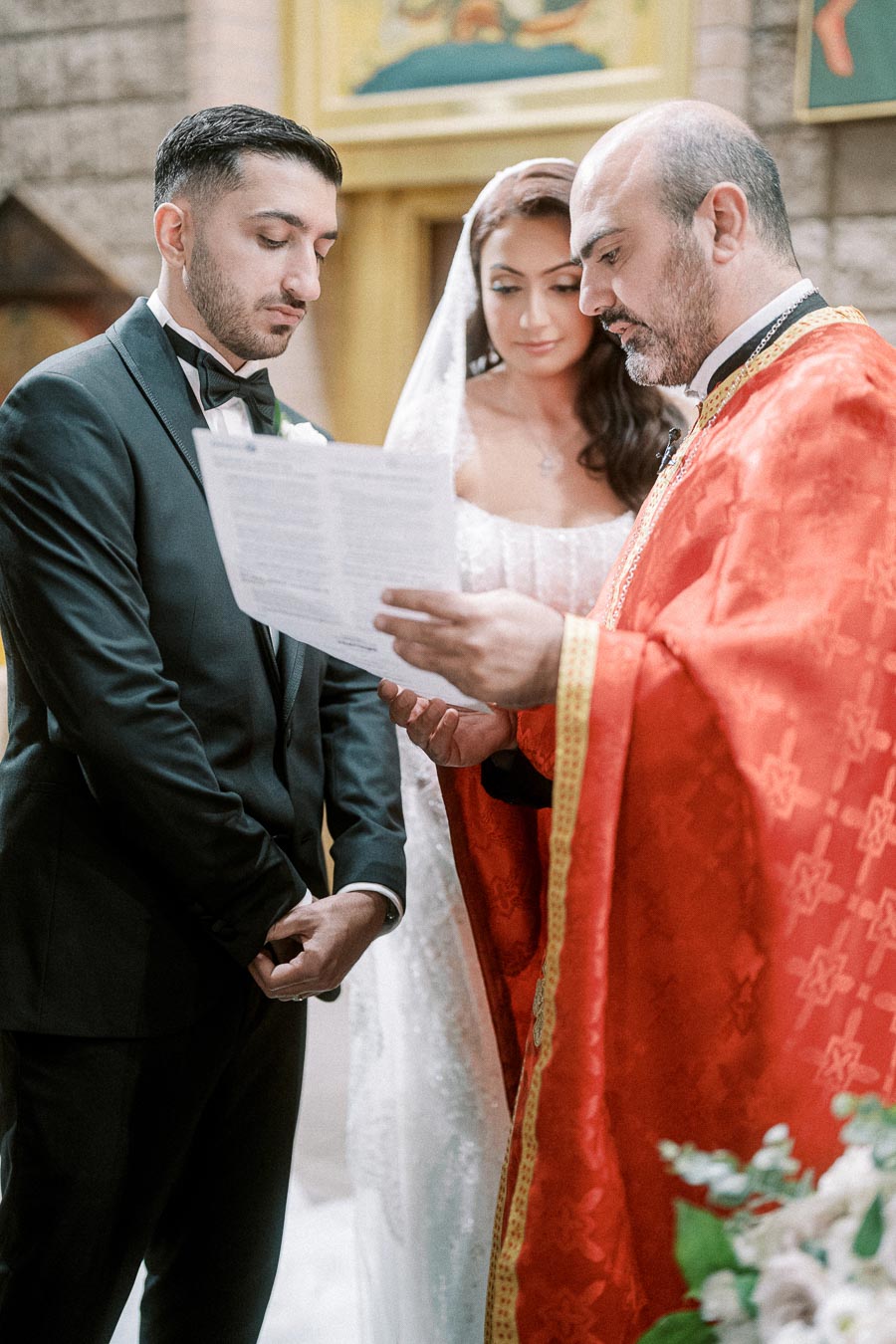 A groom and bride in wedding attire stand with a priest in a church ceremony, as the priest reads from a document. The groom