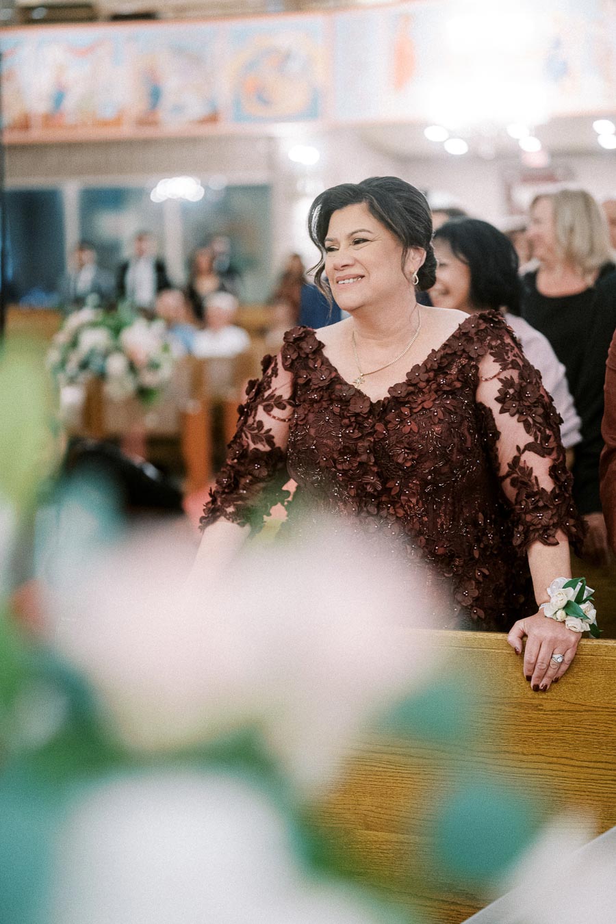 A woman in a floral embroidered dress smiles warmly during a celebration in a church, surrounded by attendees and floral