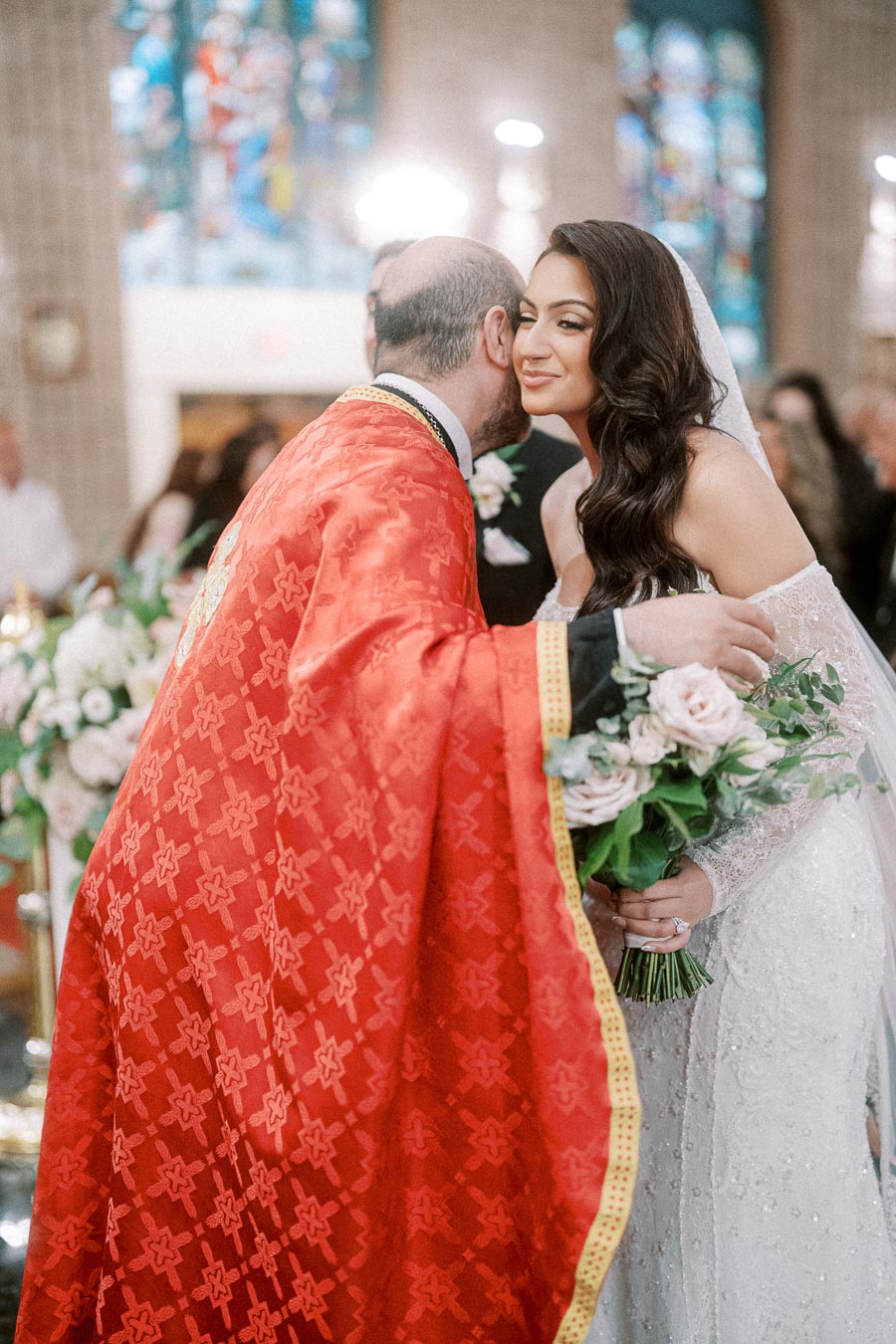 A bride in a white wedding dress holding a bouquet of pink and white roses is greeted by a priest in red ceremonial robes