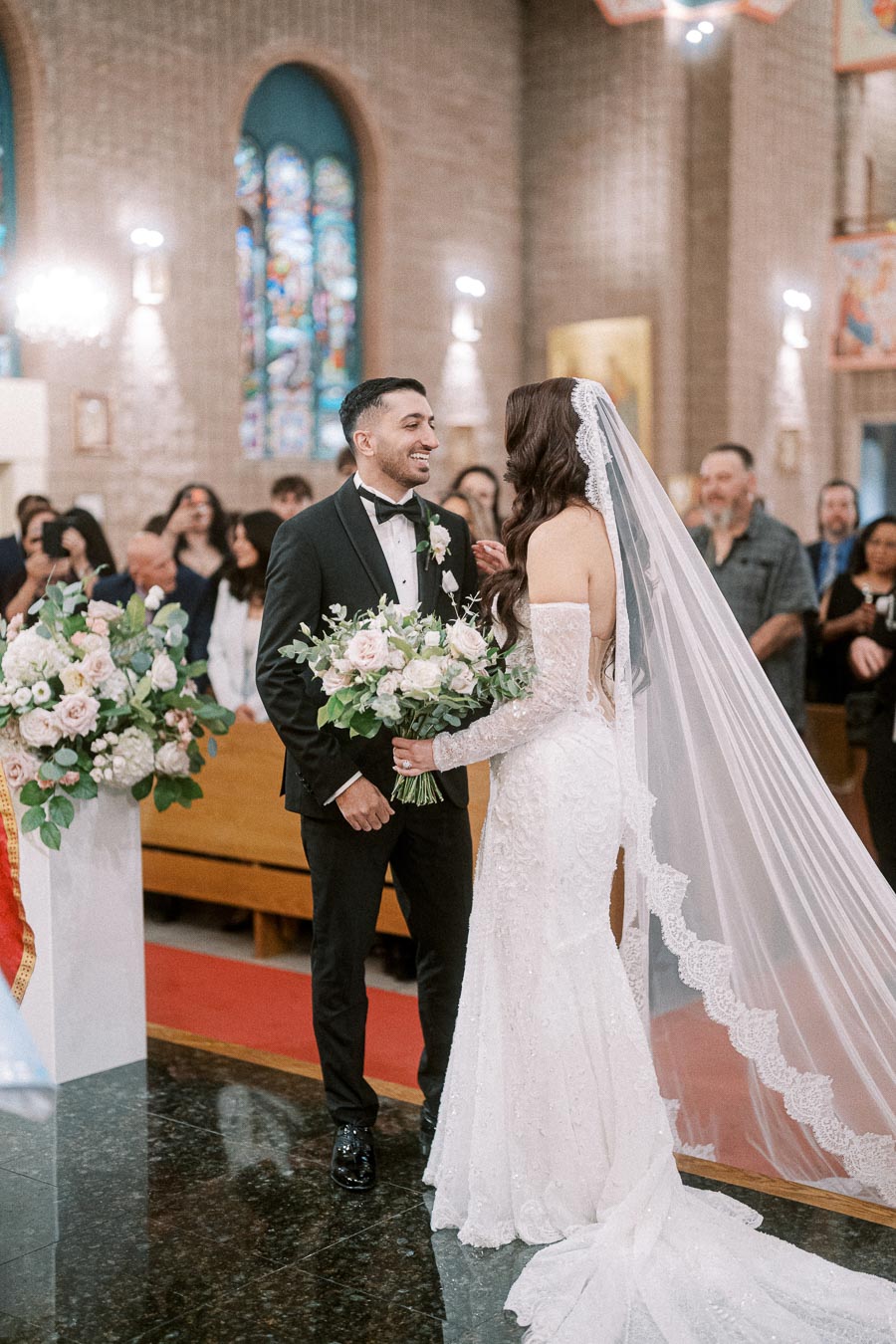 Bride and groom smiling at each other during their wedding ceremony in a beautifully decorated church, with the bride in an