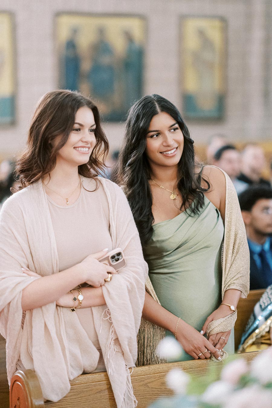 Two women smiling at a formal event, dressed elegantly with neutral and green tones, inside a beautifully decorated venue.