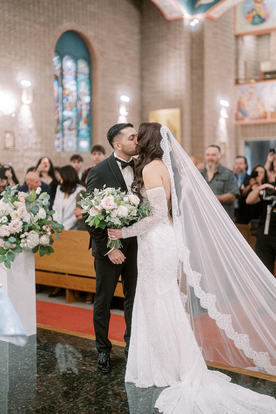 Romantic wedding ceremony with bride in elegant lace dress and long veil kissing groom in black tuxedo inside beautifully